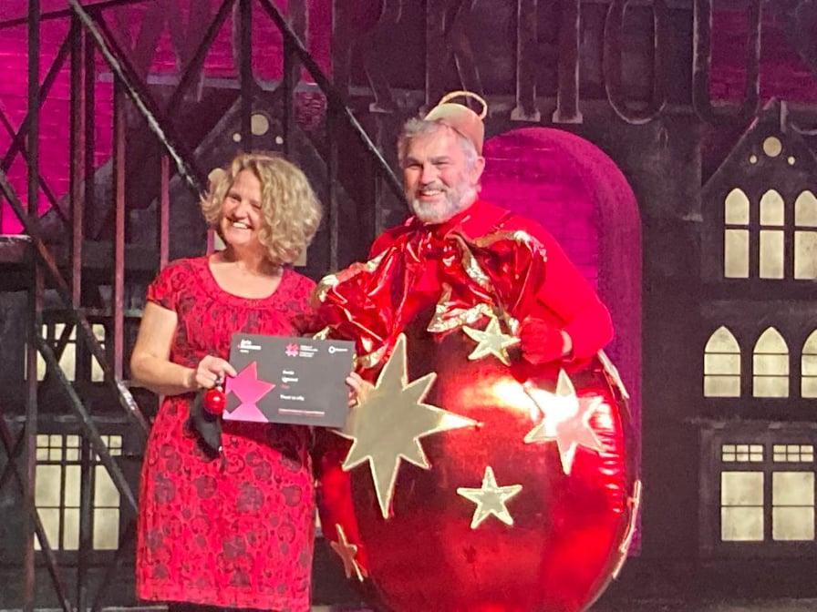 A woman with curly blonde hair wearing a red dress accepting an award from a man dressed as a red Christmas bauble